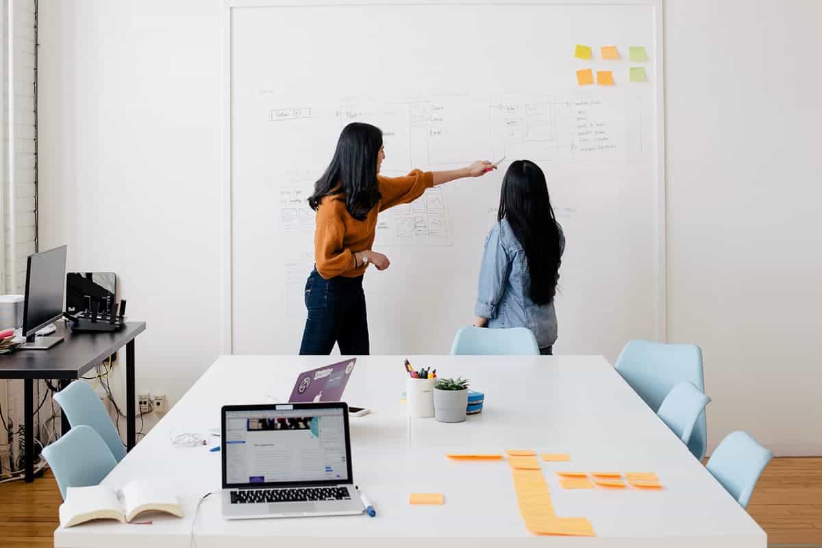 Two coworkers brainstorm at a large whiteboard with sketches and sticky notes, while a laptop and note cards sit on a conference table in a bright office.