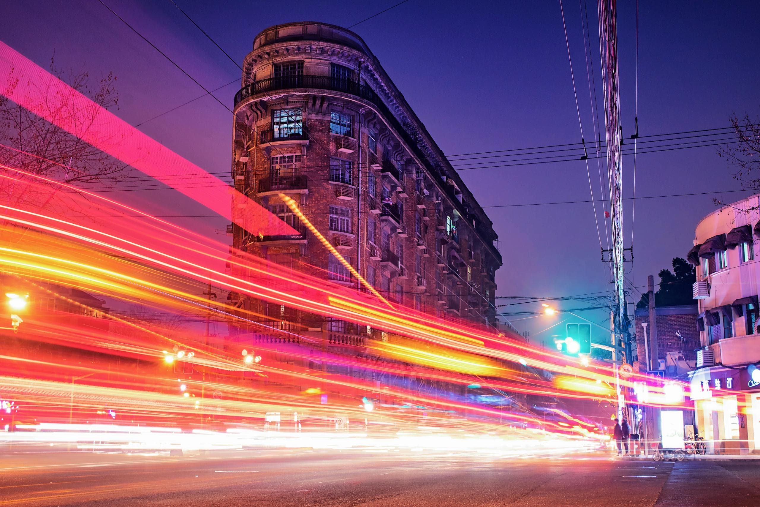 Long-exposure traffic light trails streak past a historic corner building on a city street at night.