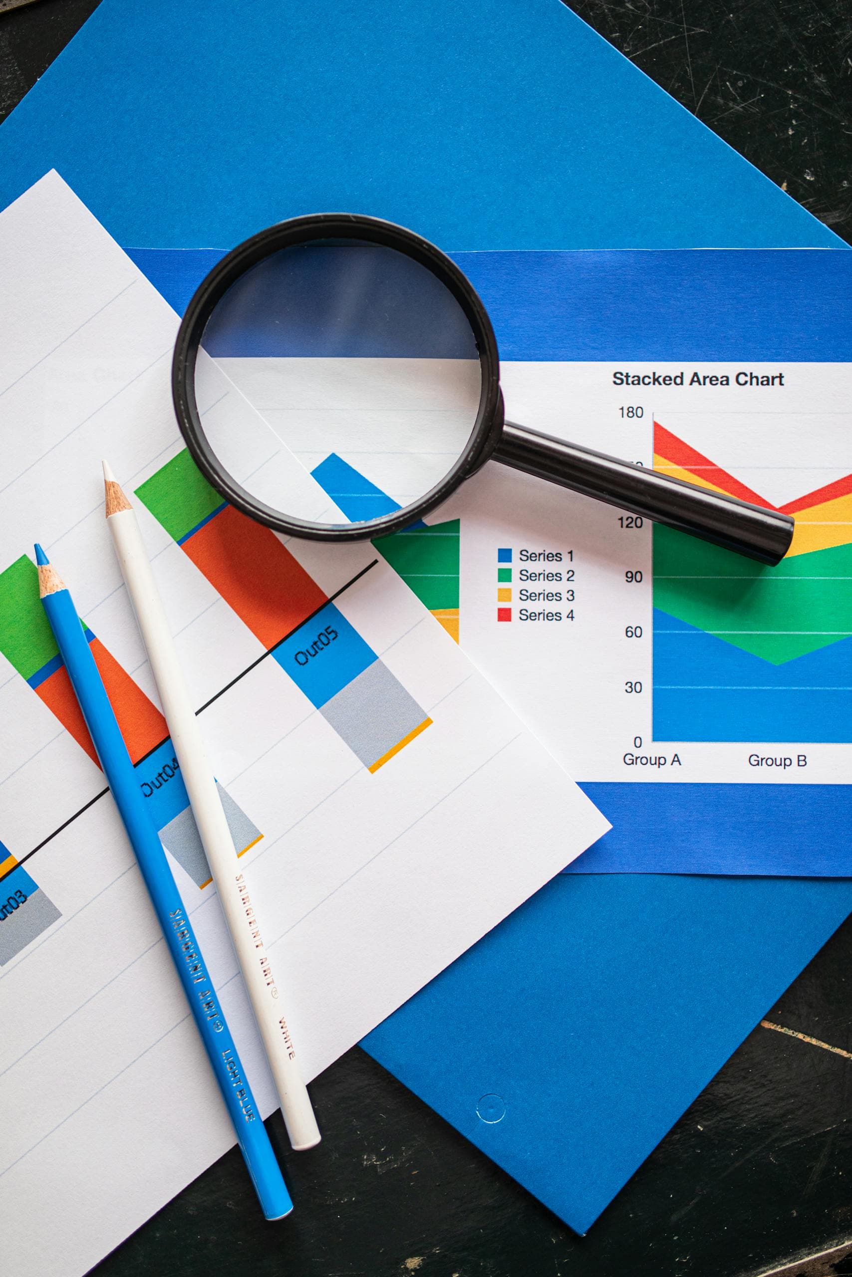 Magnifying glass over printed analytics charts and reports on a blue folder with colored pencils.