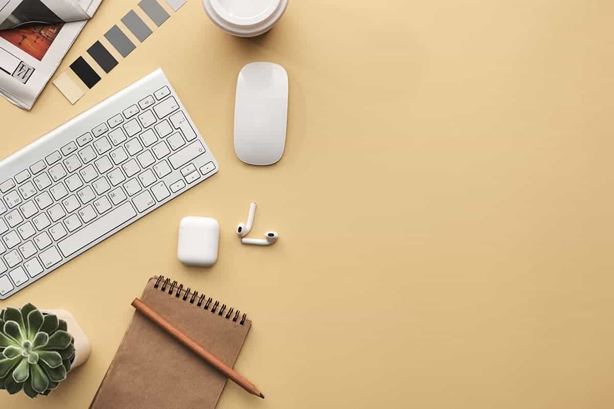Minimalist workspace flat lay with a keyboard, wireless mouse, earbuds, notebook, pencil, and small plant on a beige desk.