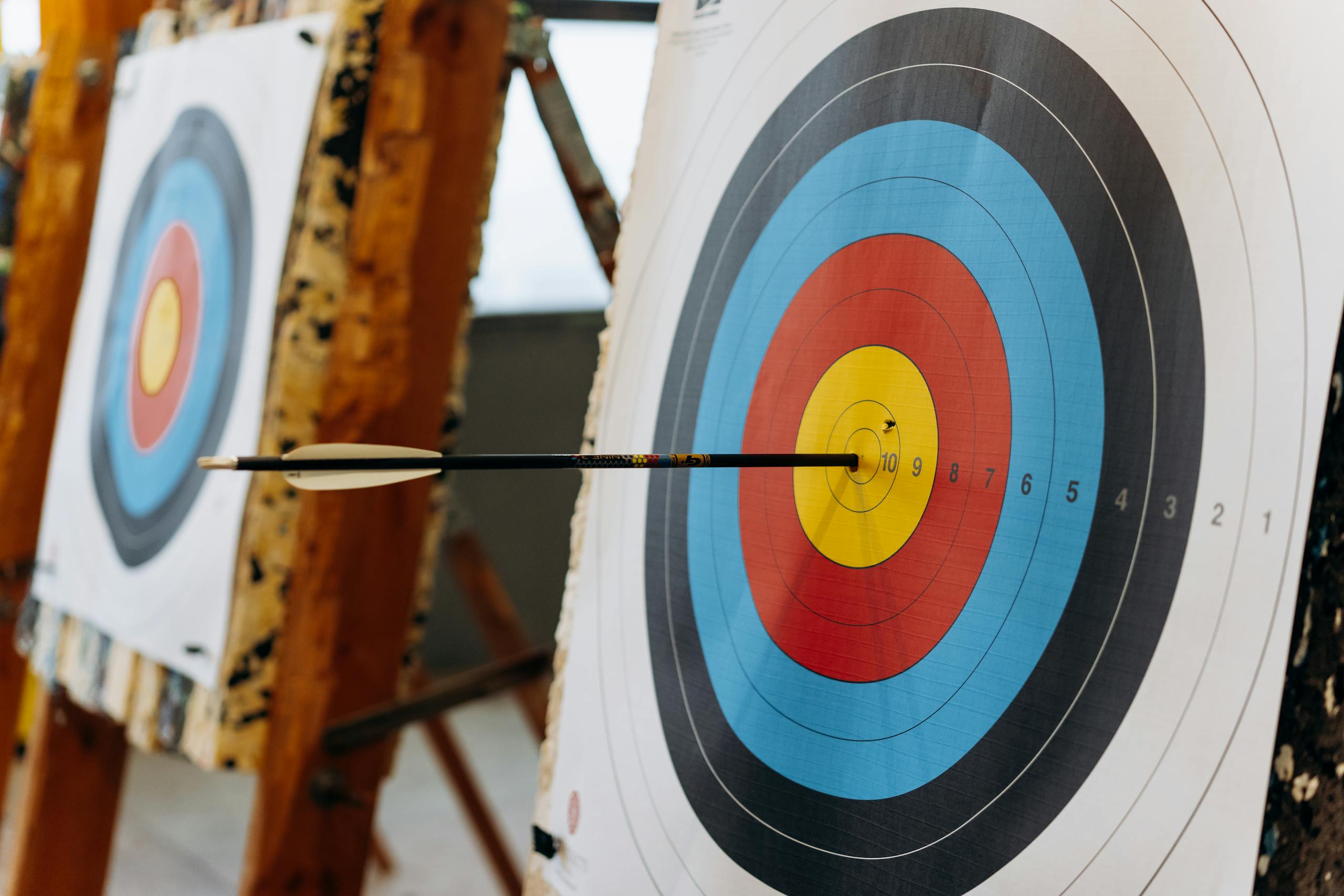 An arrow hits the bullseye on a colorful target in an indoor archery range. Archery target with an arrow lodged near the bullseye on an indoor range, with a second target blurred in the background.