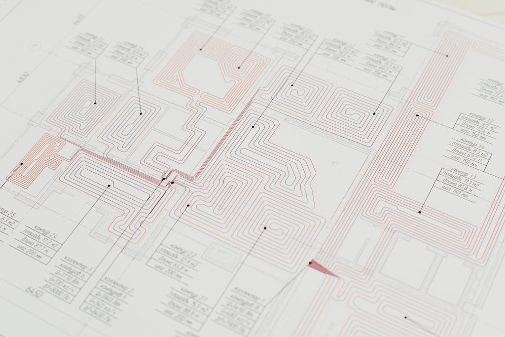 Close-up view of a detailed architectural blueprint with intricate designs and data points. Technical building plan showing a radiant floor heating pipe layout with red and blue circuit lines and labeled zones.