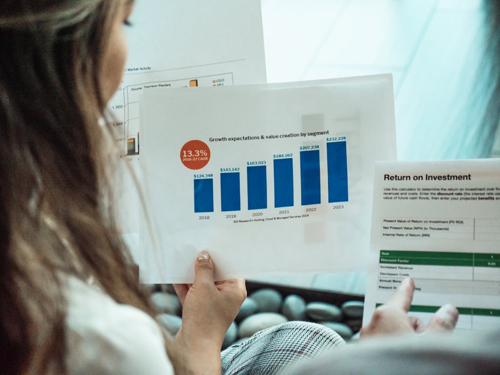 Person analyzing financial graphs and ROI reports, focusing on investment growth. Woman reviewing a printed growth forecast bar chart while another person points to a return-on-investment report.